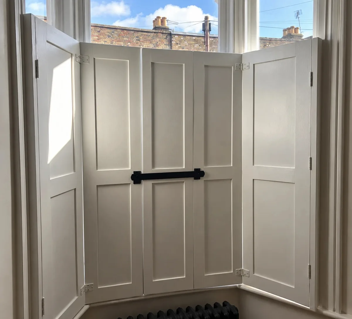 Painted panelled shutters folding across the lower half of a bay sash window, held by a black shutter bar