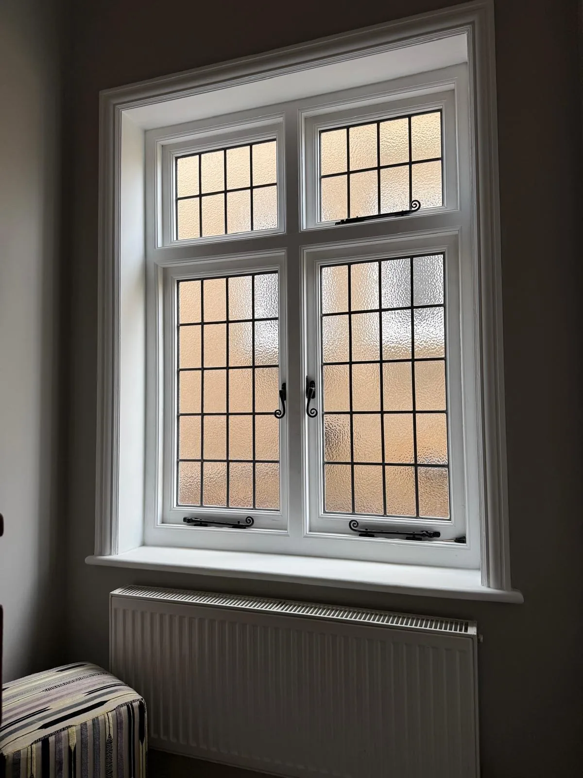A white timber casement window with leaded lights and brass fasteners above a radiator in a period room