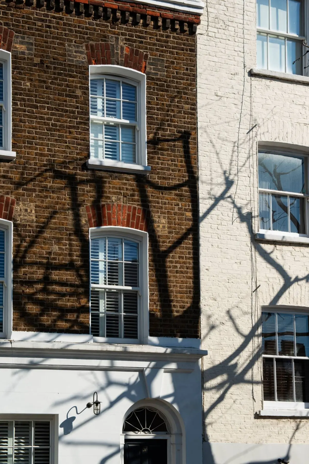 Six-over-six timber sash windows with internal shutters in a brick terrace, crossed by tree shadows, above a fanlit front door