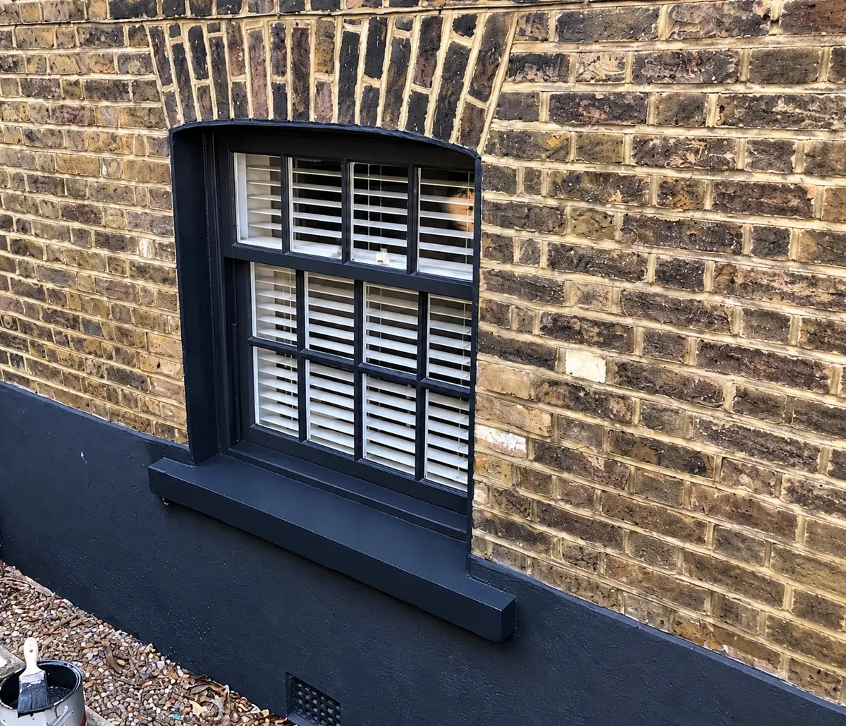 A charcoal-painted timber sash window with internal shutters, set low in weathered stock brick