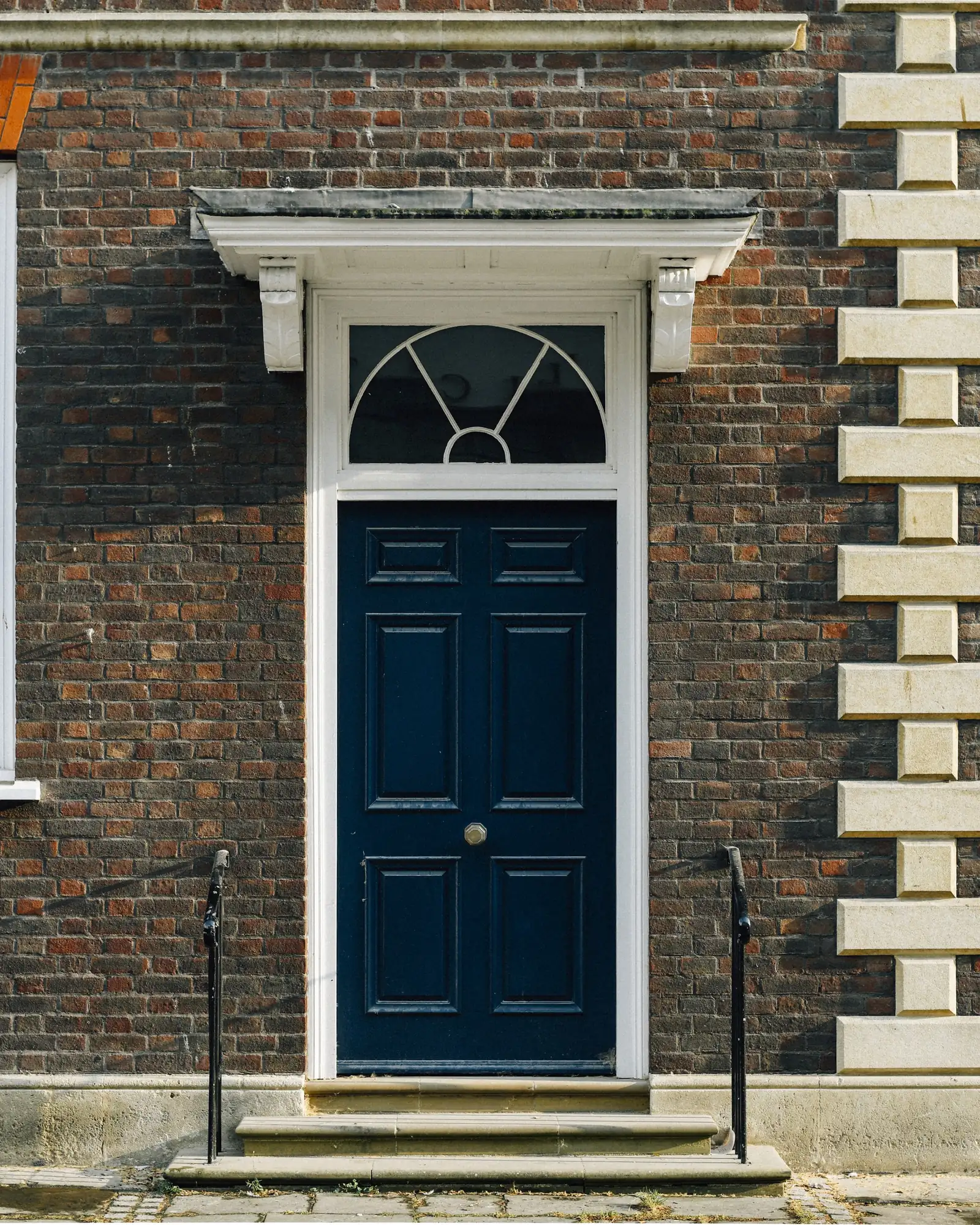 A navy six-panel front door under a radial fanlight and bracketed hood, set in brick with stone steps and black iron handrails