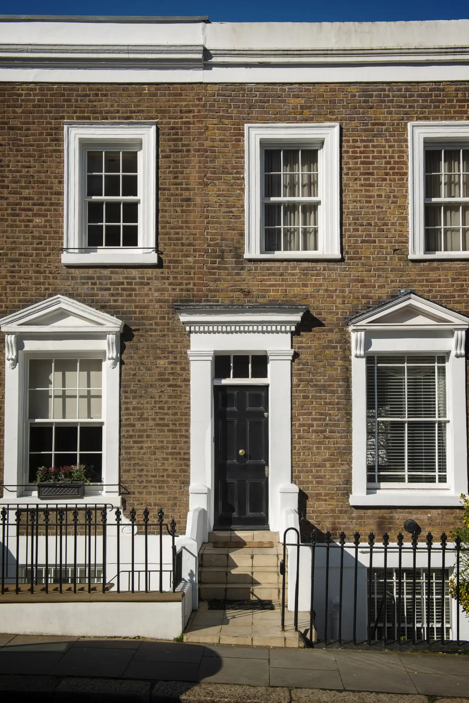 A stock-brick terraced house with white six-over-six sash windows, pedimented ground-floor surrounds and a black panelled front door behind railings