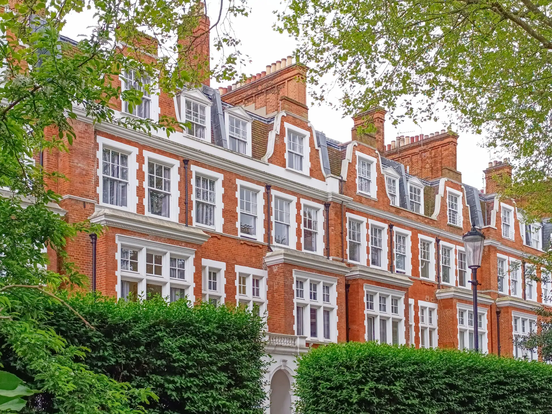 Red-brick mansion flats with white-painted sash windows, bays and Dutch gables behind a hedge