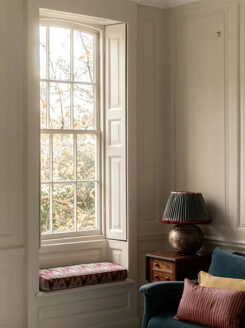 A tall timber sash window with folded shutters and a window seat in a panelled room
